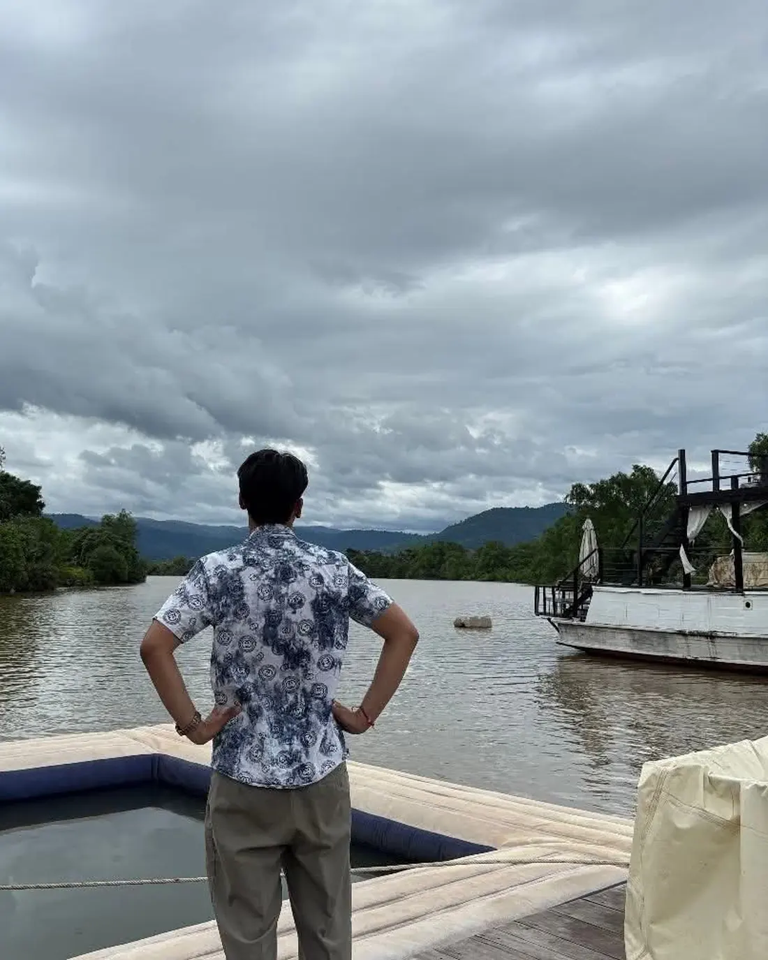 Seng Thay from behind, hands on his hips, facing a wide river and a moored boat