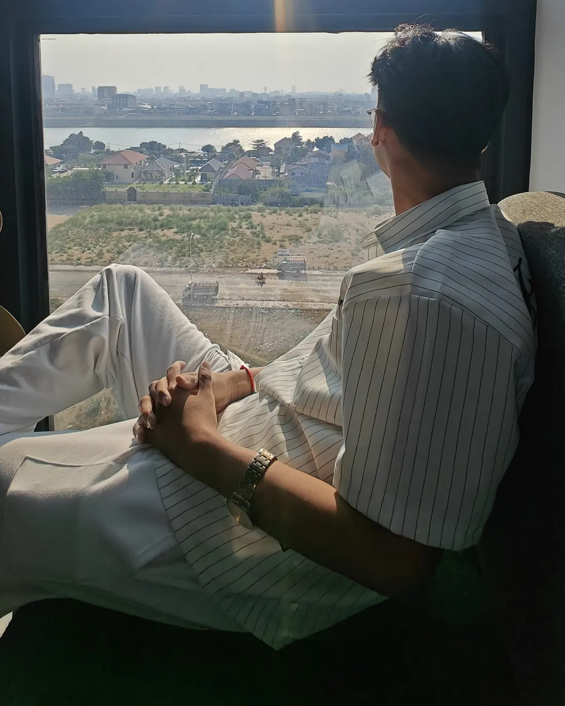 Seng Thay sitting by a tall window, looking out over a river and city skyline